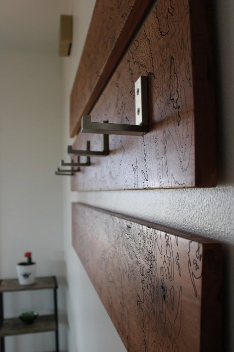 This is a three-piece work of art made of natural cherry wood, carved with topography. It is mounted on a white wall in a hallway near the home entry. The middle plank has five silver adjustable hooks that can be opened for use or closed.
